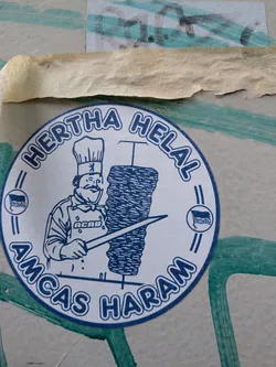 Street sticker A circular sticker printed in blue ink on a white background. It depicts a cartoon chef with a mustache, wearing a chef's hat adorned with the Berlin bear logo, and an apron with the acronym 'ACAB'. The chef is holding a long knife next to a large spit of doner kebab. The text 'HERTHA HELAL' is arched across the top, and 'AMCAS HARAM' is arched across the bottom. On the left and right sides, there are small circular icons containing the Hertha BSC football club flag.