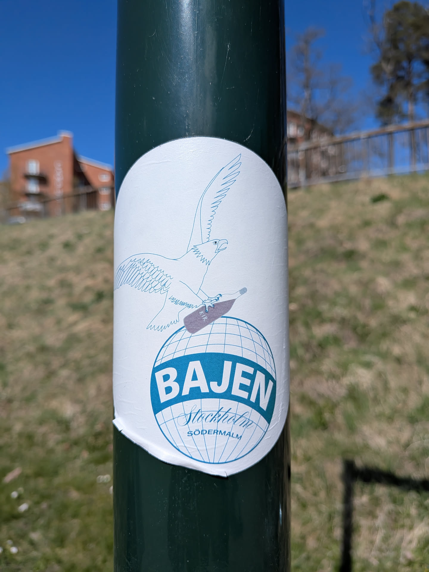 A white, roughly circular sticker featuring a blue line-art illustration of an eagle in flight, carrying a brown bottle labeled 'KIR' in its talons. Below the eagle is a blue outline of a globe with grid lines. A solid blue band crosses the globe, containing the bold white text 'BAJEN'. Beneath 'BAJEN', the words 'Stockholm' in a script font and 'SÖDERMALM' in a smaller sans-serif font are written in blue, all within the globe's design. The sticker has a slightly wrinkled and uneven bottom edge, indicating its physical application.