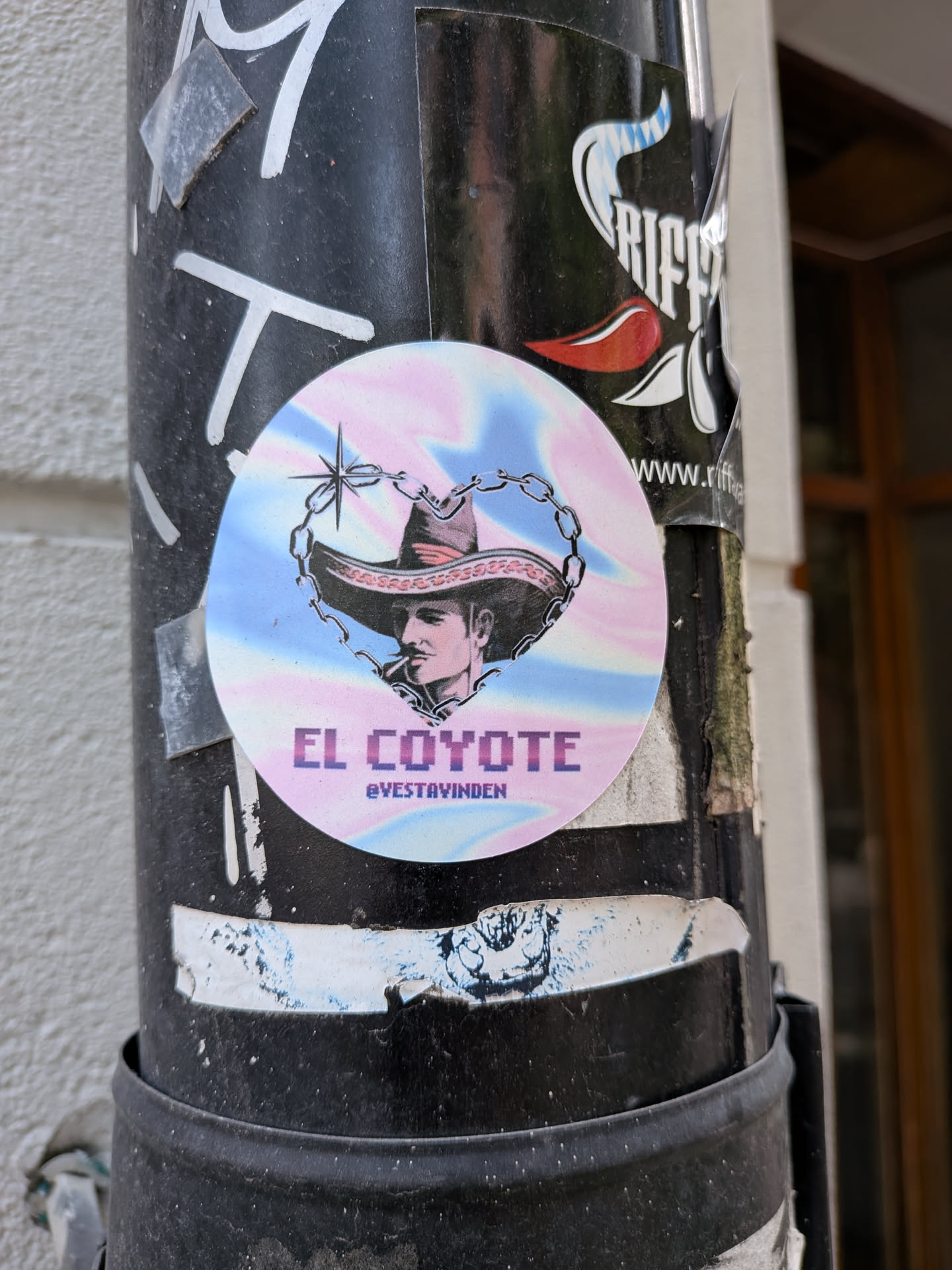 A circular sticker with a pastel pink and blue iridescent background. It depicts a black-and-white illustration of a man in a sombrero framed by a heart-shaped chain. Below the image, the text 'EL COYOTE' and the handle '@VESTAVINDEN' are displayed.