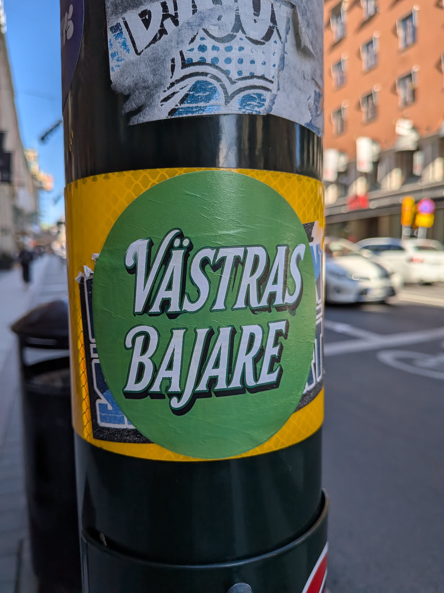 Circular green sticker featuring stylized white text 'VÄSTRAS BAJARE' with black drop shadows, affixed to a yellow reflective band on a dark pole.