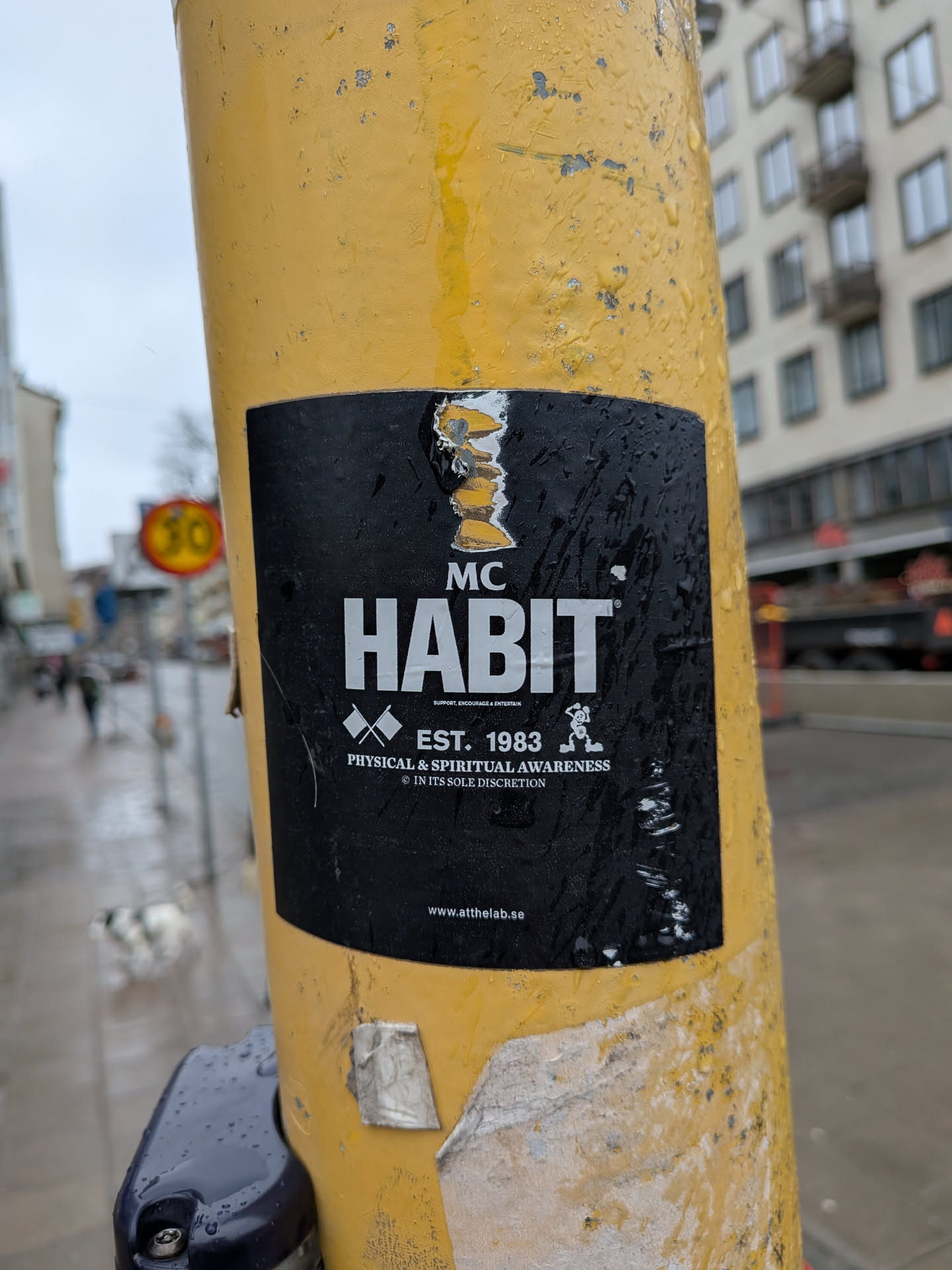 A rectangular sticker with a matte black background and white text and graphics, adhered to a textured yellow pole. At the top, 'MC' is in a smaller font above 'HABIT' in large, bold, sans-serif letters. Below 'HABIT' is the tagline 'SUPPORT. ENCOURAGE & ENTERTAIN'. Further down, two white crossed flags are positioned to the left of 'EST. 1983'. Below this, 'PHYSICAL & SPIRITUAL AWARENESS' is written, followed by '© IN ITS SOLE DISCRETION'. To the right of the establishment date and awareness statement, a small, stylized white cartoon character, possibly a monkey or similar figure, is depicted in an active, dynamic pose. At the very bottom, the website 'www.atthelab.se' is printed. The sticker is wet with water droplets and shows signs of wear, with slight peeling at its top and bottom edges.