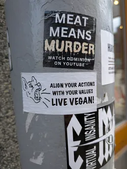 Street sticker A rectangular white sticker featuring a bold black and white design. On the left side, there's a stylized, somewhat aggressive illustration of a dog or wolf-like head with its mouth open, appearing to bark or howl, indicated by radiating lines. To the right, the text "ALIGN YOUR ACTIONS WITH YOUR VALUES" is stacked above "LIVE VEGAN!", all in a strong, sans-serif black font. The overall aesthetic is clean and impactful, conveying a direct message.