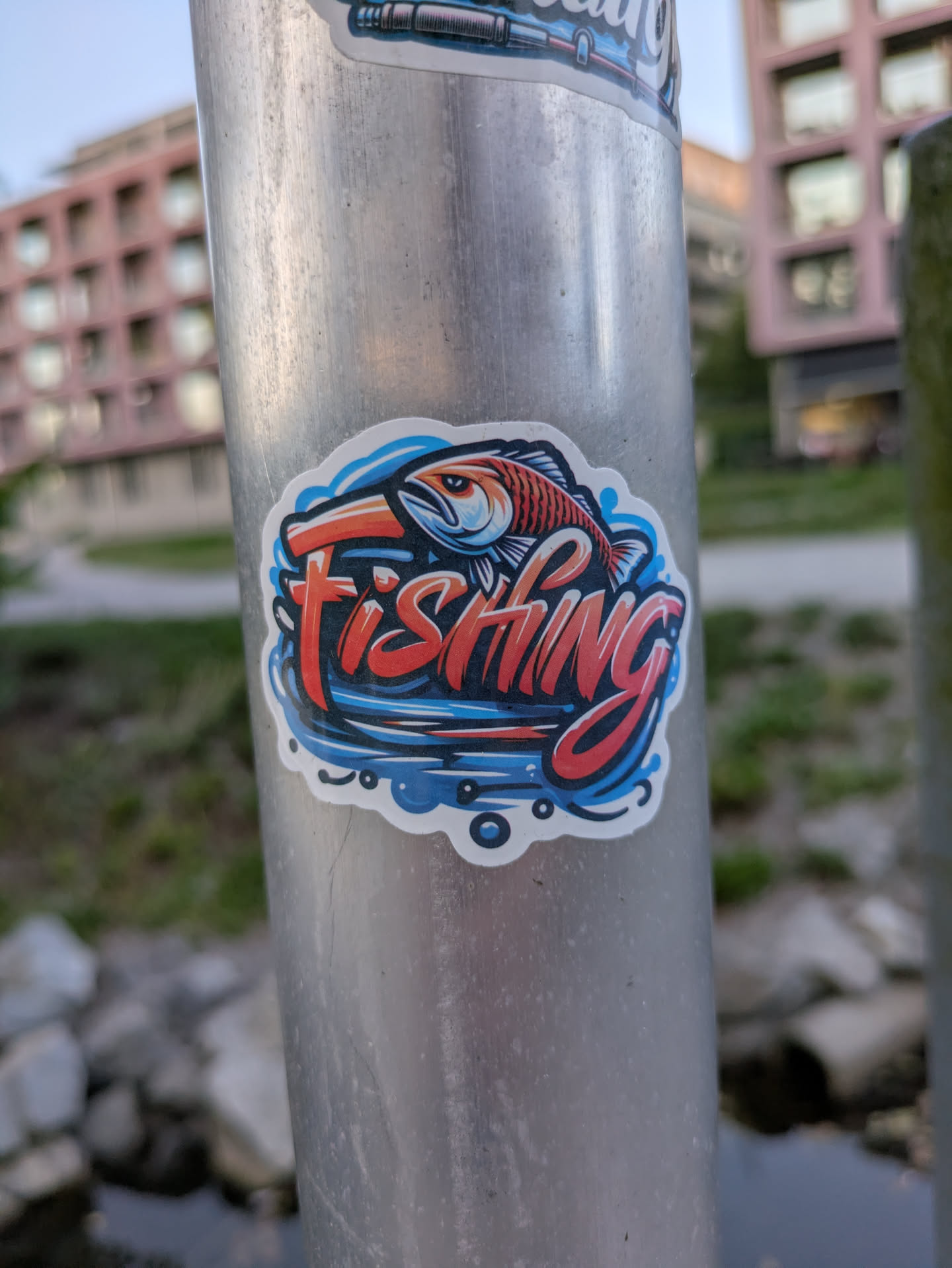 A dynamic sticker featuring a stylized fish leaping from water, with the word 'FISHING' prominently displayed. The fish is depicted in shades of orange and red with black outlines and white highlights, appearing to be a bass or similar game fish. Below and around the fish, stylized water splashes are rendered in various shades of blue, from light to dark, with white foam details. The word 'FISHING' is in bold, red, block letters with a darker outline and white highlights, giving it a three-dimensional effect. The entire design is set against a dark background within the sticker and is bordered by a clean white outline, making it stand out against the metallic pole.