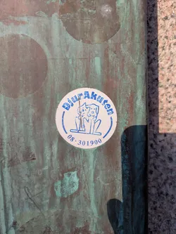 Street sticker This is a round sticker for Djurakuten, a Swedish animal hospital, placed on a mottled, potentially copper, surface. The sticker is white with blue text and graphics. It includes the name of the hospital and a phone number.
