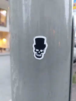 A sticker of a skull wearing a top hat is affixed to a light gray pole. The sticker is simple, black and white, with a minimalist design.