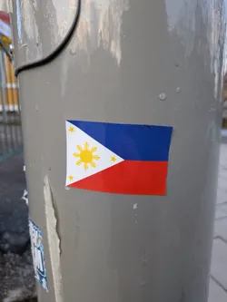 Street sticker The sticker depicts the national flag of the Philippines. It features two horizontal bands of equal size, with a royal blue band on top and a scarlet red band on the bottom. A white equilateral triangle is positioned at the hoist side. Centered within the white triangle is a golden-yellow sun with eight primary rays, each representing a province. In each corner of the white triangle, there is a smaller golden-yellow five-pointed star. The sticker shows minor wear and slight peeling along its top edge.