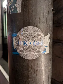 Street sticker The sticker is circular and features a highly symmetrical, intricate geometric pattern in shades of gray and white. The pattern resembles a stylized network of lines and diamonds. In the center, the text 'IFK GBG' is prominently displayed in a bold, blue, sans-serif font. A small blue diamond shape is positioned between 'IFK' and 'GBG'. The sticker appears to be adhered to a rough, textured surface, possibly a metal pole, with some surrounding peeling paper.