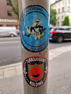 A round sticker with a blue background shows a cartoon bird wearing a sailor's hat and holding two beer steins. The text "Turbojugend Hässleho" is visible around the image.  Another sticker below it is black, red and green, showing a shield-like emblem with anchors and the text "Turbojugend Kolding".