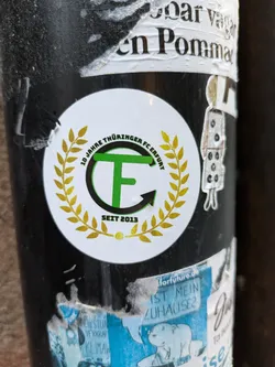 Street sticker A round sticker commemorating 10 years of Thüringer FCE Erfurt (since 2013). It features a green and gold logo with the letters 'T' and 'F' inside a circular design encircled by a laurel wreath. 