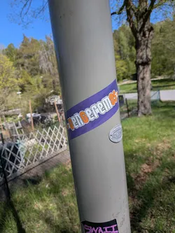 Street sticker The sticker is a rectangular band, oriented diagonally across the pole. It features a solid purple background. The word 'VENCEREMOS' is prominently displayed in a bold, uppercase font, colored orange with a distinct white outline. The text has a distressed or grunge effect, giving it a worn appearance. A small orange star precedes the word 'VENCEREMOS'.