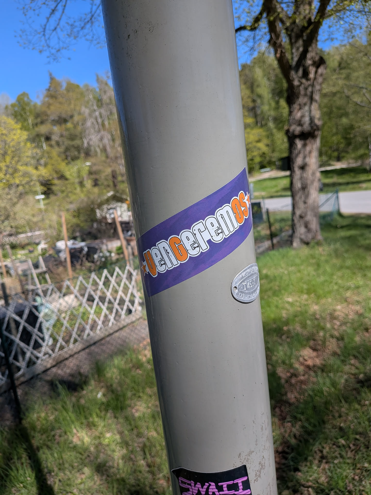 The sticker is a rectangular band, oriented diagonally across the pole. It features a solid purple background. The word 'VENCEREMOS' is prominently displayed in a bold, uppercase font, colored orange with a distinct white outline. The text has a distressed or grunge effect, giving it a worn appearance. A small orange star precedes the word 'VENCEREMOS'.