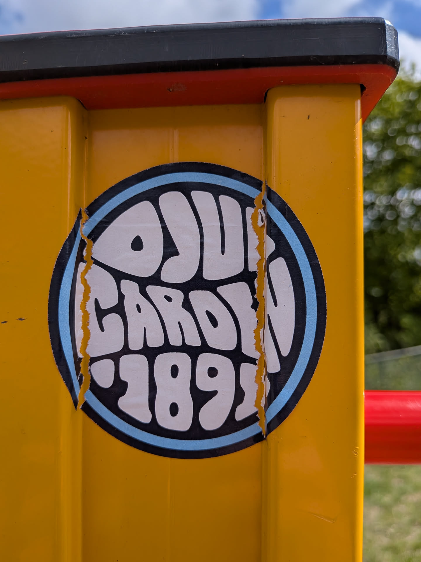 A circular sticker featuring bold, white, bubbly lettering that reads 'OYUN GARDEN '89'. The text is set against a black background, encircled by a light blue border. The words 'OYUN' and 'GARDEN' are stacked, with ''89' at the bottom, all following the curve of the circle. The sticker shows signs of wear, with jagged tears on both the left and right sides, revealing the yellow surface of the pole beneath.