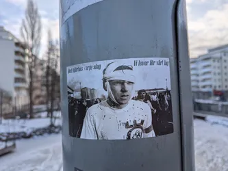 Street sticker A rectangular, horizontally oriented sticker featuring a black and white photograph. The central figure is a young man, likely a football player, with a bandaged head covering his eyes and forehead. He wears a light-colored t-shirt splattered with dark marks, possibly blood or mud. On his right arm and chest, a circular emblem with a stylized 'AIK' logo is visible. In the blurred background, a crowd of spectators and some industrial-looking structures can be seen. The overall aesthetic is gritty and evokes a sense of historical struggle or intense dedication.