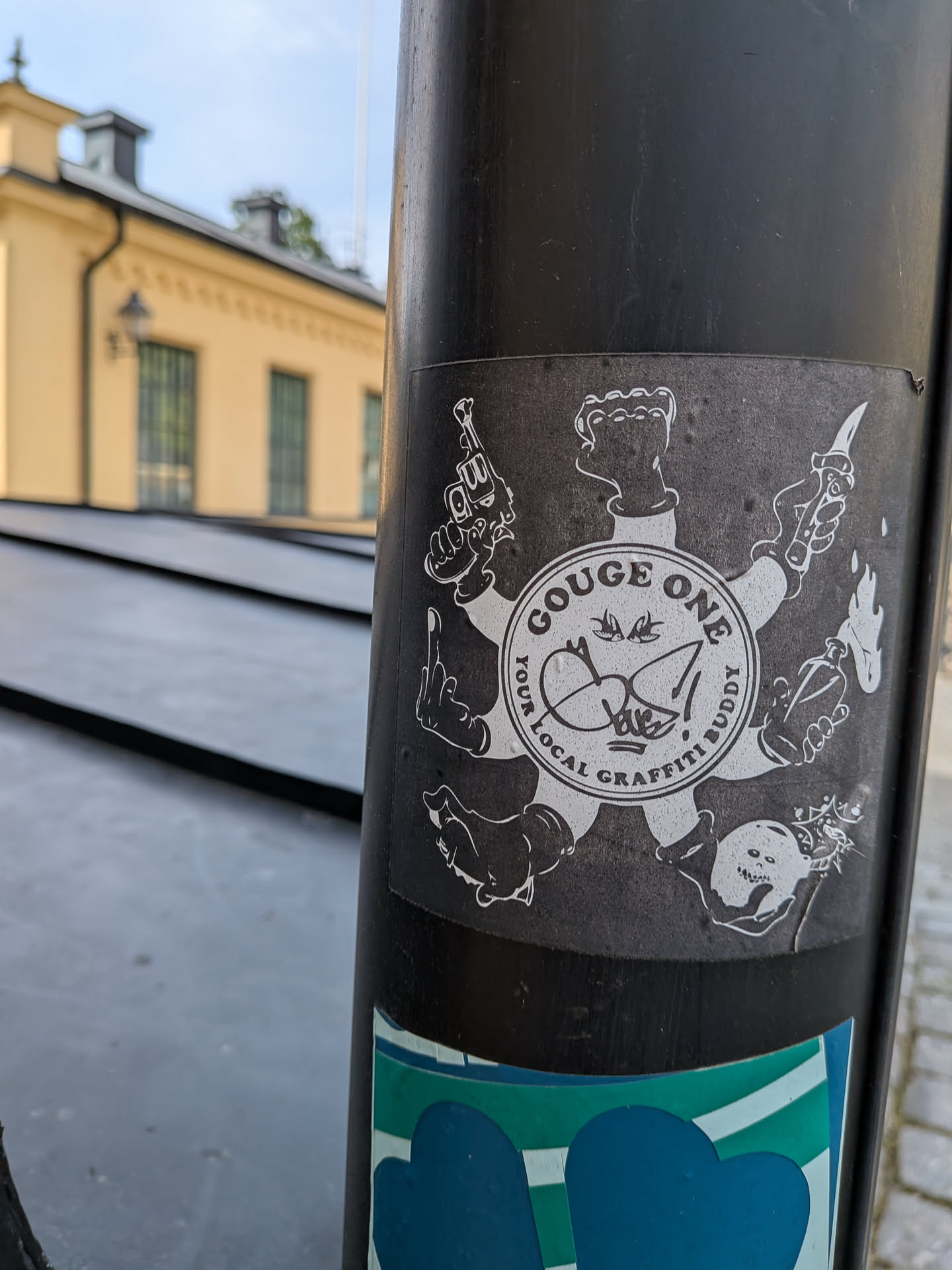 A square black sticker with a white circular design. It depicts a central emblem with seven radiating arms, each holding an object or making a gesture: a revolver, a middle finger, a boot, a bomb, a Molotov cocktail, a knife, and a clenched fist. The inner circle features two swallows, a graffiti tag, and the text 'GOUGE ONE YOUR LOCAL GRAFFITI BUDDY'.