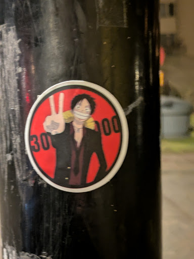 This is a round sticker featuring an illustration inspired by the One Piece character Monkey D. Luffy. The character is wearing a suit and making a peace sign, with a bounty of "300.000" written on the red background behind him. The sticker is adhered to a black pole.