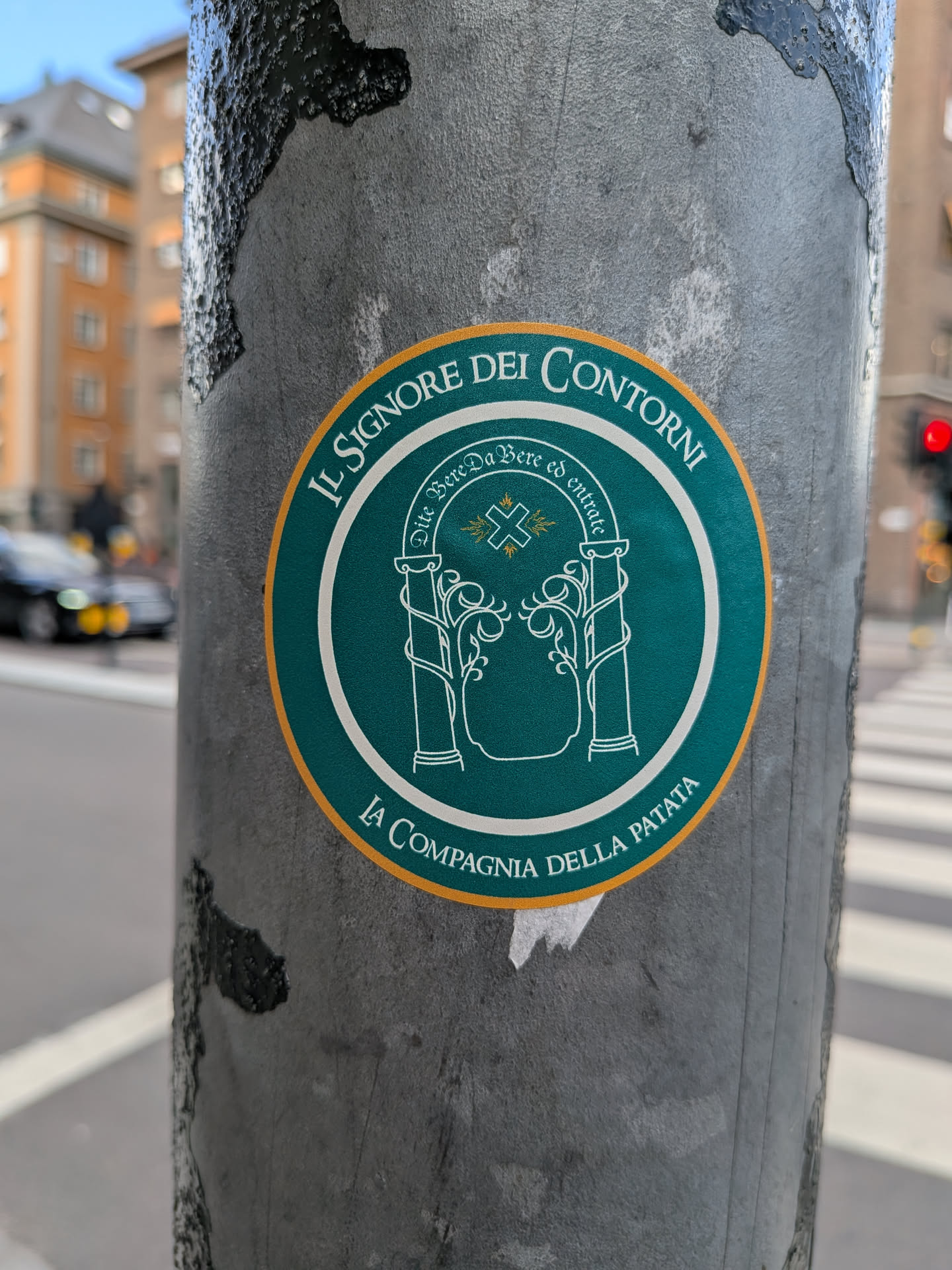 Circular teal sticker with a yellow border. It depicts a white line-art illustration of the Doors of Durin. Italian parody text reads 'IL SIGNORE DEI CONTORNI' and 'LA COMPAGNIA DELLA PATATA'.