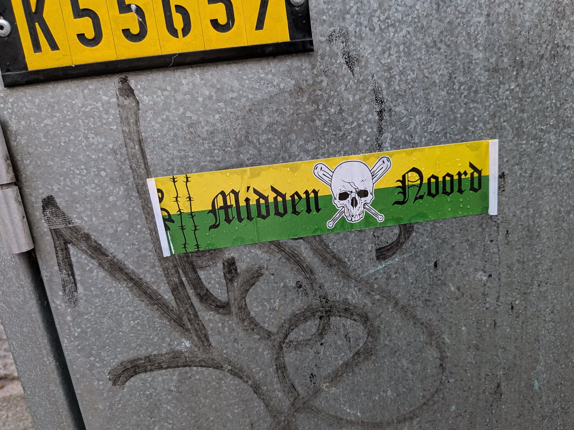 Street sticker The sticker is rectangular and horizontally oriented. It features a yellow band at the top and a green band at the bottom, separated by a thin black line. On the left side of the sticker, there are vertical black lines resembling barbed wire. In the center, a white skull with crossed baseball bats behind it is depicted. To the right of the skull, the word 'Midden' is written in a gothic-style font in black, followed by the word 'Noord' in the same font and color. The sticker appears to have water droplets on its surface, suggesting it has been exposed to the elements.