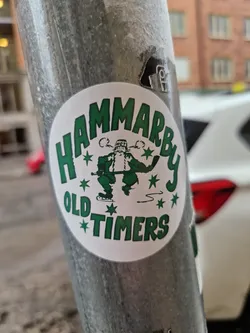 Street sticker A round sticker featuring a cartoon illustration of a hockey player. The text 'Hammarby Old Timers' is written in a green vintage-style font around the illustration. The sticker is affixed to a grey metal pole.