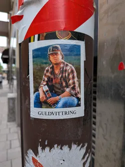 Street sticker A rectangular sticker with a white border showing a man in a camo cap, glasses, and a plaid shirt sitting outdoors. He holds a Molotow Burner 600 spray can. The Swedish word 'GULDVITTRING' is printed at the bottom.