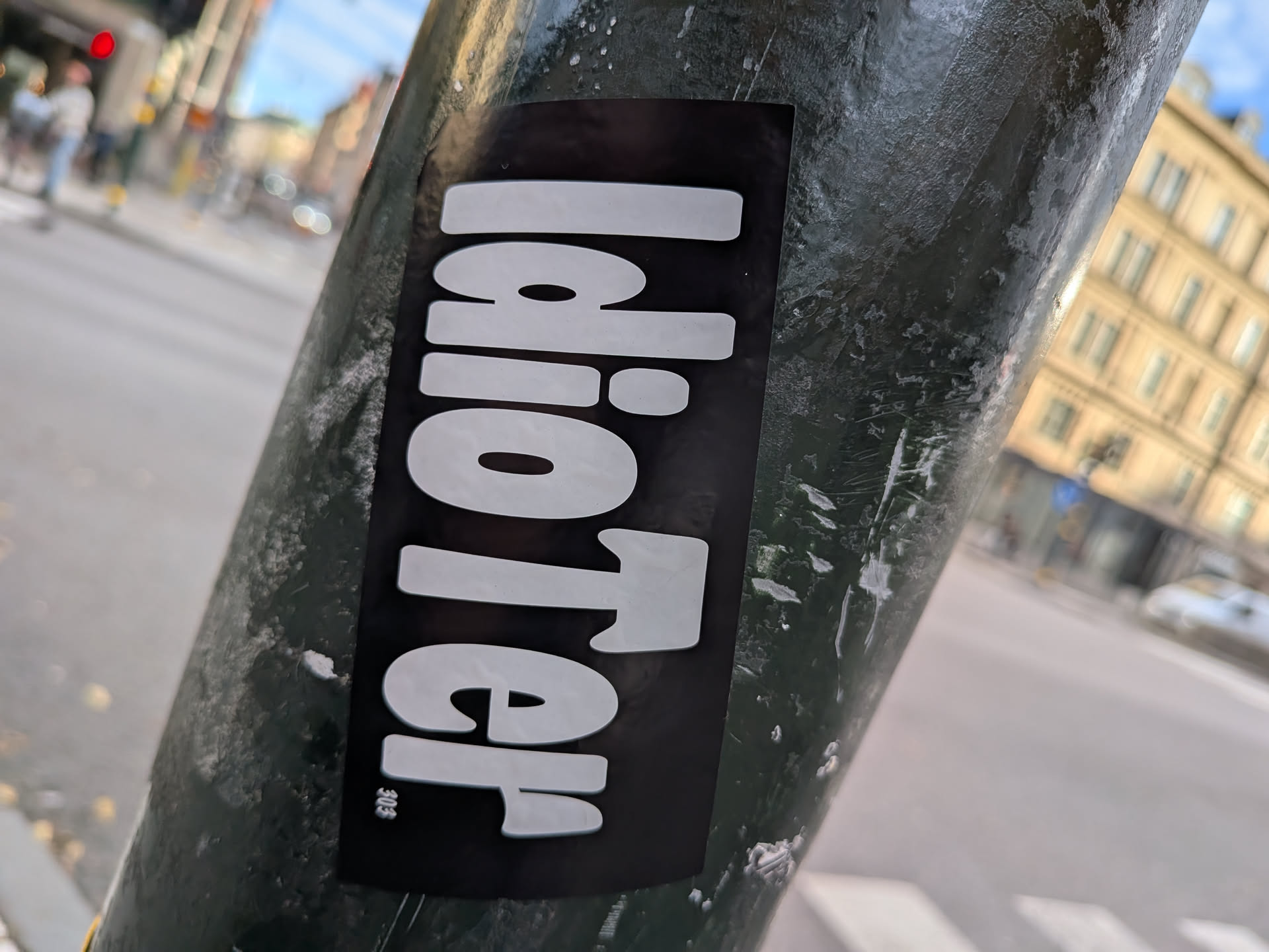 The sticker is rectangular with rounded corners and features the word 'Idioter' in large, bold, white sans-serif letters against a solid black background. The letters have a subtle shadow effect, giving them a slightly raised appearance. In the bottom left corner of the black background, there is a small, stylized logo or symbol that appears to be '305' in a very small font. The sticker is affixed to a dark, textured pole, with some wear and tear visible on the pole's surface.