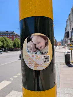 Street sticker The sticker is an oval-shaped, worn, and peeling decal affixed to a black and yellow striped pole. The upper portion displays a color photograph of two children: an older girl with reddish-brown hair smiling down at a baby with light hair, who appears to be sleeping. The lower portion of the sticker has a pale yellow background with partially obscured red text and a black-and-white QR code on the right. The text is heavily damaged and peeling, revealing the black pole beneath. The overall appearance suggests a long-standing public display.