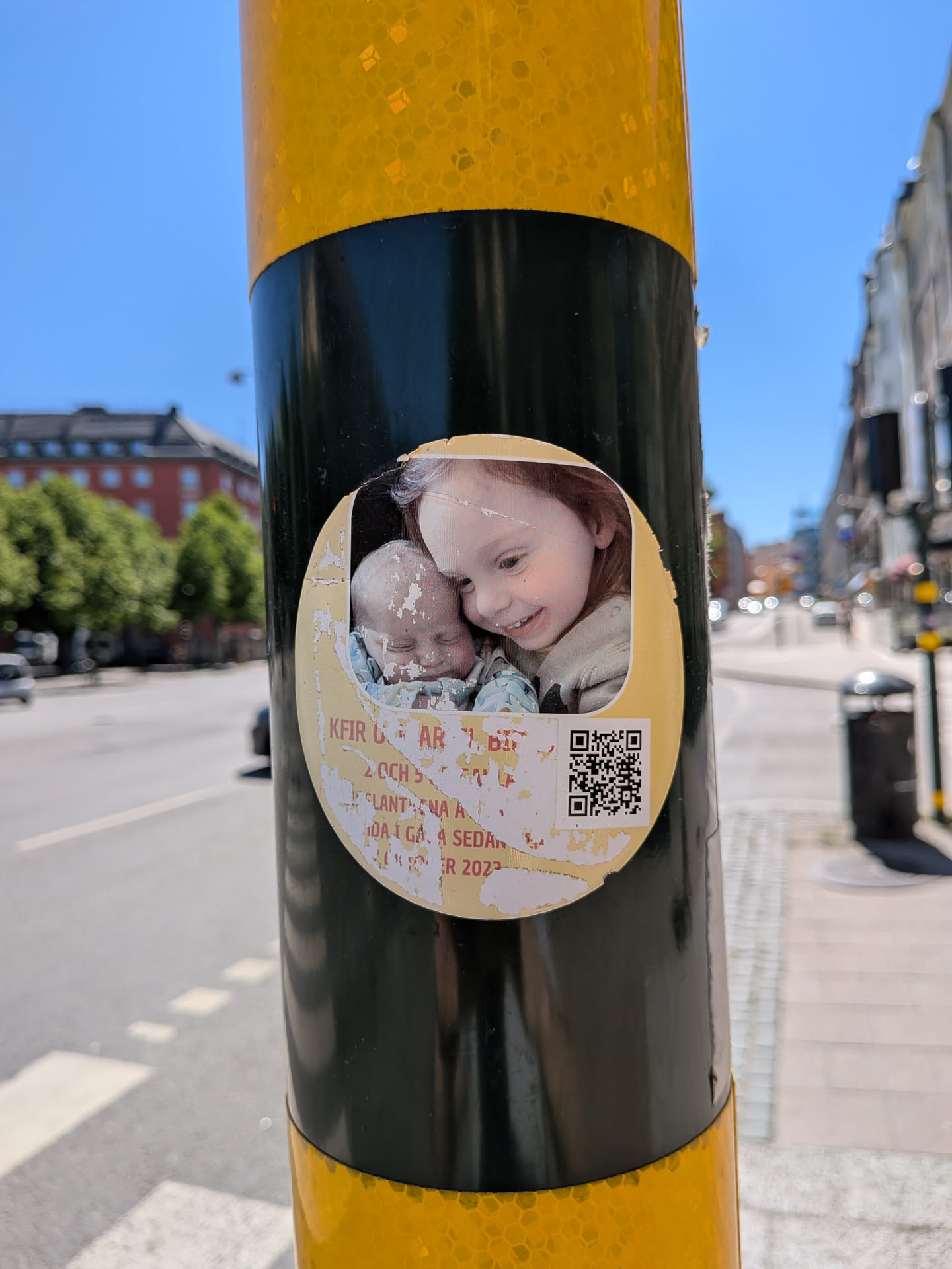 The sticker is an oval-shaped, worn, and peeling decal affixed to a black and yellow striped pole. The upper portion displays a color photograph of two children: an older girl with reddish-brown hair smiling down at a baby with light hair, who appears to be sleeping. The lower portion of the sticker has a pale yellow background with partially obscured red text and a black-and-white QR code on the right. The text is heavily damaged and peeling, revealing the black pole beneath. The overall appearance suggests a long-standing public display.