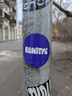 Street sticker A round, purple sticker with the word \"Kehitys\" written in a stylized, bubbly white font. The sticker is affixed to a metal pole, amongst other stickers.