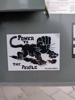 Street sticker The sticker features a bold, black and white graphic design. The central image is a stylized black panther-like creature in a crouching stance, with its left front paw transformed into a large, clenched fist raised in a gesture of power or solidarity. The panther has sharp claws on its other visible paws and prominent whiskers. Above the panther's back, the text 'POWER TO' is written in a bold, slightly distressed font. Below its belly, 'THE PEOPLE' is written in the same style. In the bottom right corner, the phrase 'DELICIOUS BRAINS' is present in a smaller, more decorative script font. The sticker has a white background with all design elements rendered in solid black, creating a high-contrast and impactful visual.
