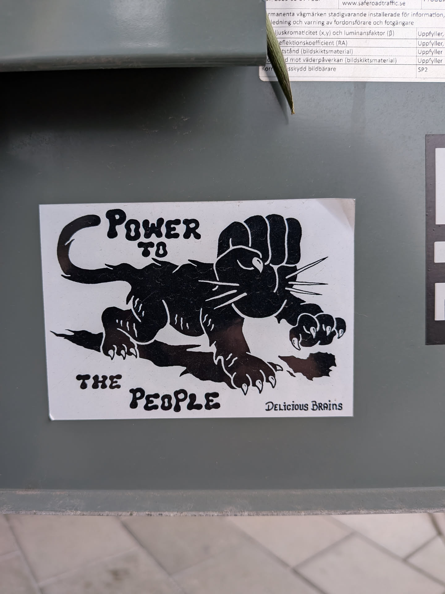 The sticker features a bold, black and white graphic design. The central image is a stylized black panther-like creature in a crouching stance, with its left front paw transformed into a large, clenched fist raised in a gesture of power or solidarity. The panther has sharp claws on its other visible paws and prominent whiskers. Above the panther's back, the text 'POWER TO' is written in a bold, slightly distressed font. Below its belly, 'THE PEOPLE' is written in the same style. In the bottom right corner, the phrase 'DELICIOUS BRAINS' is present in a smaller, more decorative script font. The sticker has a white background with all design elements rendered in solid black, creating a high-contrast and impactful visual.