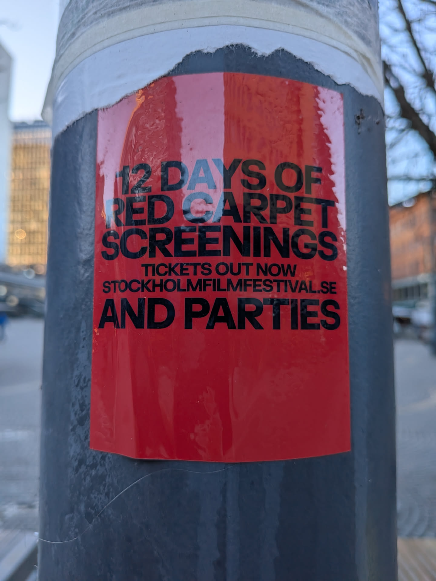 A vibrant red rectangular sticker adhered to a dark grey pole. The sticker features bold black sans-serif text. The top portion reads "12 DAYS OF RED CARPET SCREENINGS", followed by smaller text "TICKETS OUT NOW STOCKHOLMFILMFESTIVAL.SE", and concludes with "AND PARTIES" in large black letters at the bottom. The sticker shows signs of wear, with some wrinkles and slight peeling at its top edge, revealing a white layer underneath. The surface has a somewhat textured, slightly glossy appearance.