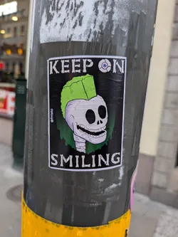 Street sticker The sticker features a cartoonish skull with a bright green mohawk, a wide, toothy grin, and empty black eye sockets. One eye socket contains a stylized eyeball with a pink iris and blue pupil. The skull is depicted with a light purple hue and is outlined in black. The background is black, with green dripping elements behind the skull. The text 'KEEP ON' is at the top and 'SMILING' is at the bottom, both in white, stylized, bone-like lettering with a black outline. A small '@fntstc' handle is visible vertically on the left side of the sticker.