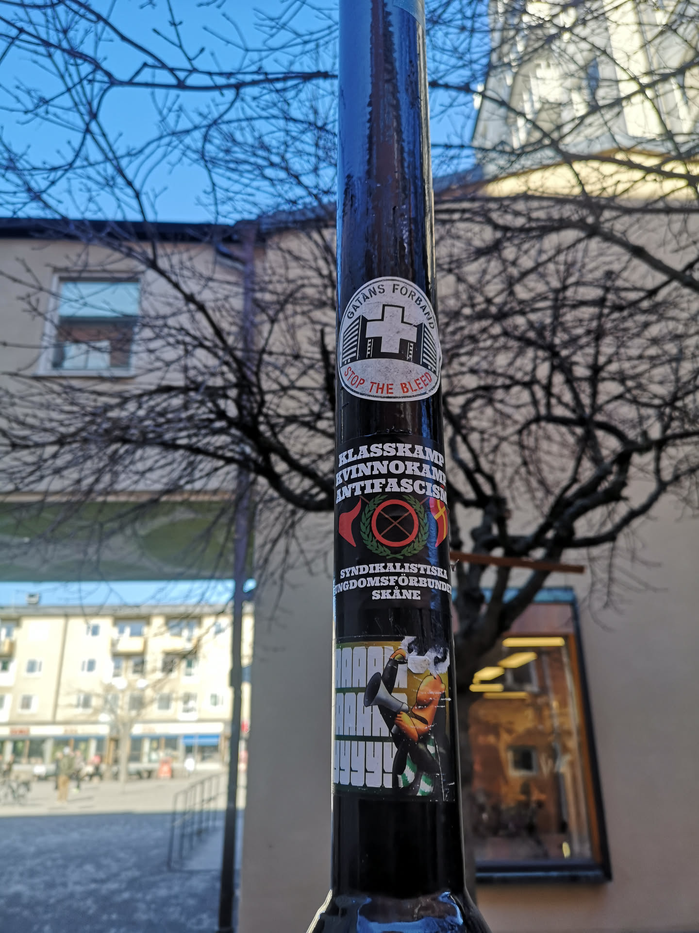 The image displays three distinct stickers vertically arranged on a dark, glossy pole. The topmost sticker is an oval white decal with black text 'GATANS FÖRBUND' arcing at the top and 'STOP THE BLEED' in red text arcing at the bottom. Its center features a stylized white cross symbol flanked by black building silhouettes. Below it is a rectangular black sticker with prominent white text: 'KLASSKAMP', 'KVINNOKAMP', and 'ANTIFASCISM' stacked vertically. This sticker also includes a central circular emblem with a red border, a green wreath, and a black circle with a red diagonal cross (a common anti-fascist symbol), flanked by a red flag on the left and a yellow/red flag on the right. Further down on this sticker, it reads 'SYNDIKALISTISKA UNGDOMSFÖRBUNDET SKÅNE'. The bottom sticker is a rectangular piece featuring a cartoonish, orange-skinned character with a green and white striped garment, holding a black megaphone. Large, blocky white text 'HAAARGHH!' with a yellow outline is prominently displayed next to the character. The stickers show signs of wear, slight peeling, and some dirt.