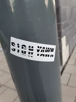 Street sticker A white sticker with black text that says "SIGH YAWN" is affixed to a gray pole. The sticker is slightly damaged.