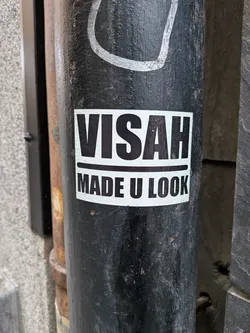 Street sticker The sticker is rectangular with a white background and black text. The top line of text reads 'VISAH' in large, bold, sans-serif capital letters. Below this, separated by a thin black line, is the text 'MADE U LOOK', also in bold, sans-serif capital letters. The sticker appears to be applied to a black, cylindrical object, possibly a pole, which shows signs of wear and tear.