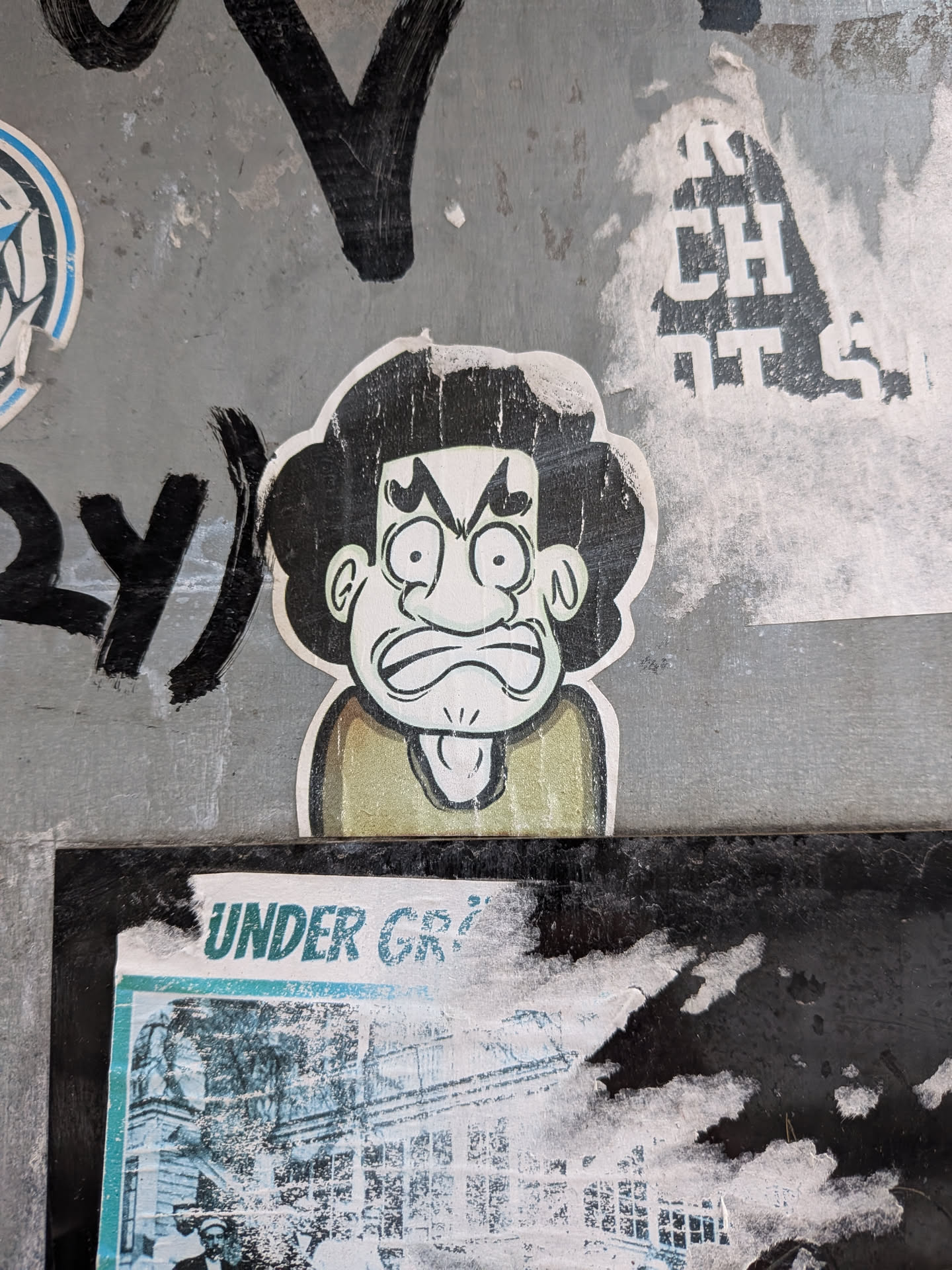 The sticker features a cartoon character's head with a highly expressive, angry, or exasperated facial expression. The character has large, dark, curly or bushy hair, outlined in white. Its face is pale, possibly light green, with wide, bulging eyes, furrowed brows, and a scrunched-up nose. The mouth is open, showing a visible tongue, and is drawn with a wavy line, emphasizing frustration. A hint of a collar or shirt in a muted olive green or khaki color is visible below the head. The entire image is outlined with a white border, suggesting it's a cut-out. The style is reminiscent of a hand-drawn doodle or comic art.