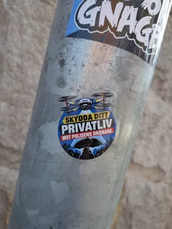 Street sticker Circular sticker depicting a police drone shining a spotlight on a silhouetted person holding an umbrella. Features Swedish text warning against drone surveillance over a dark city background.