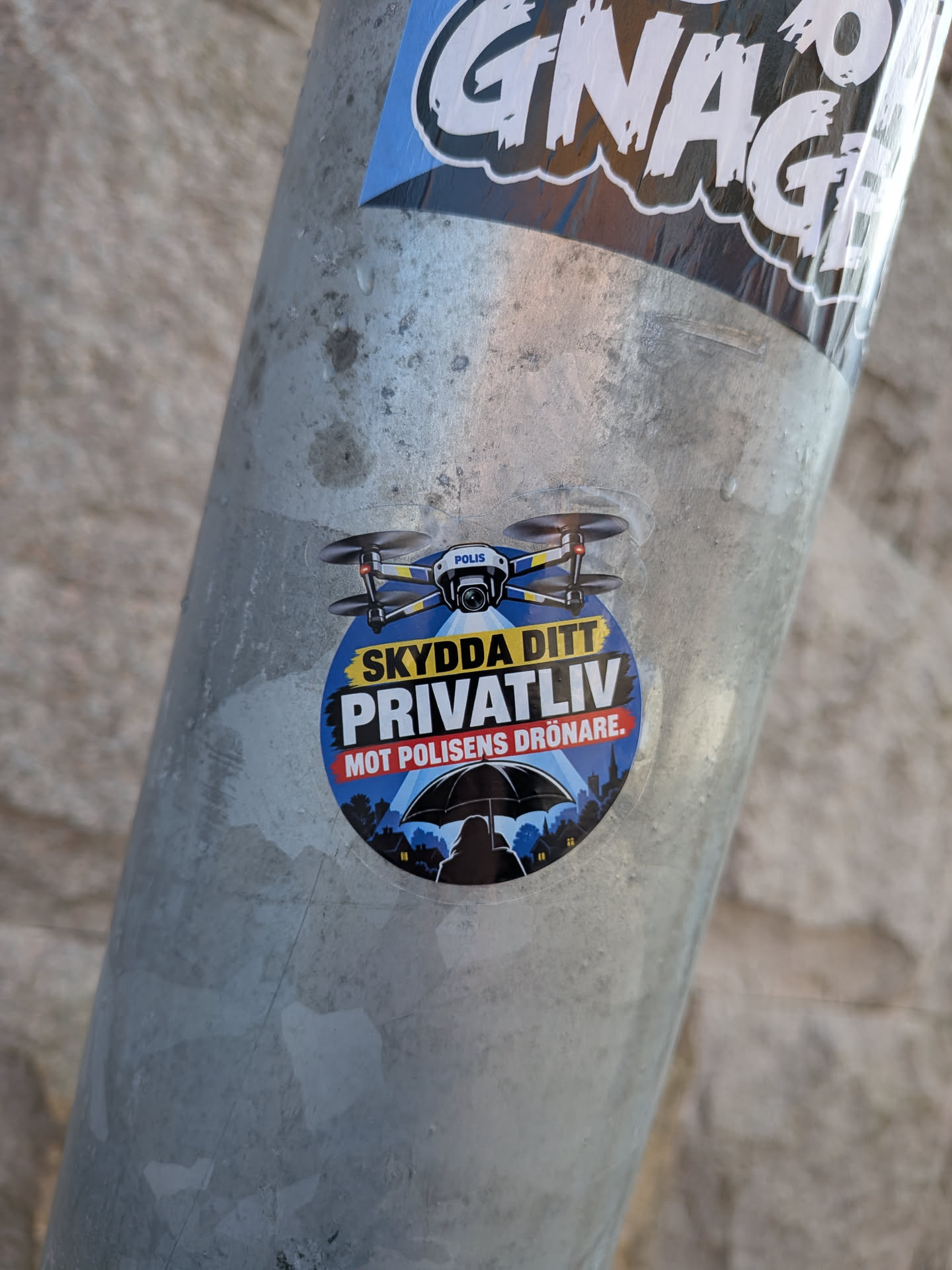 Circular sticker depicting a police drone shining a spotlight on a silhouetted person holding an umbrella. Features Swedish text warning against drone surveillance over a dark city background.
