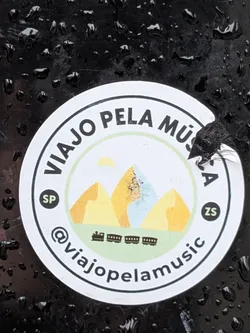 Street sticker The sticker is circular with a white background and a black border. It features a stylized landscape with three mountains in shades of yellow and light blue, under a yellow sun. Below the mountains, there is a depiction of a train with several carriages. The text 'VIAJO PELA MÚSICA' is written in a circular arc around the top of the sticker. On the left side, within a small dark circle, is 'SP', and on the right side, within a similar circle, is 'ZS'. The bottom of the sticker has the text '@viajopelamusic' in a circular arc. The sticker appears to be slightly torn on the right edge.