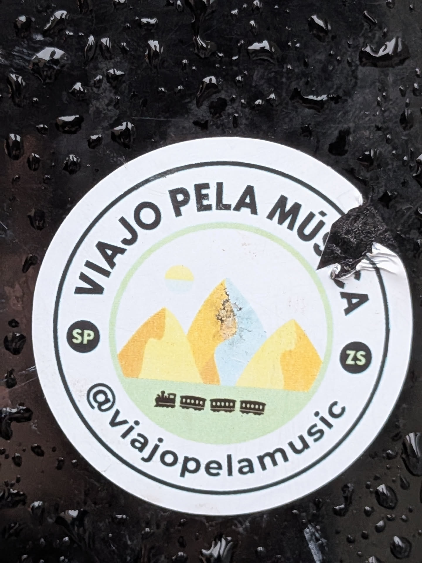 Street sticker The sticker is circular with a white background and a black border. It features a stylized landscape with three mountains in shades of yellow and light blue, under a yellow sun. Below the mountains, there is a depiction of a train with several carriages. The text 'VIAJO PELA MÚSICA' is written in a circular arc around the top of the sticker. On the left side, within a small dark circle, is 'SP', and on the right side, within a similar circle, is 'ZS'. The bottom of the sticker has the text '@viajopelamusic' in a circular arc. The sticker appears to be slightly torn on the right edge.