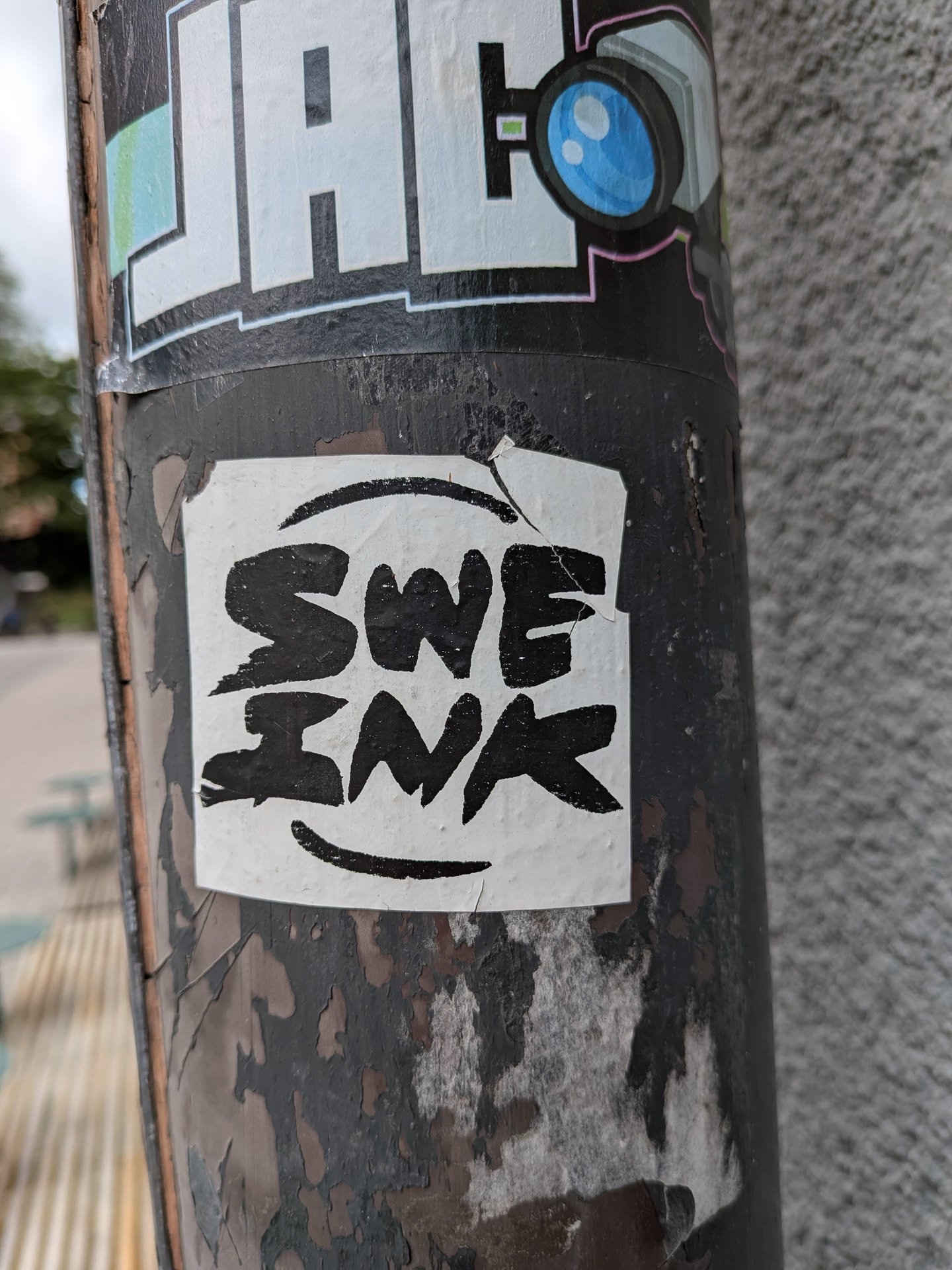 The sticker is square with slightly irregular edges, predominantly white with bold black lettering. The text reads 'SWE INK' in a thick, stylized font. A large, curved black line is positioned above the text, and a smaller, curved line is beneath it, resembling a smile. The sticker appears to be adhered to a dark, weathered pole, with peeling paint visible around its edges.
