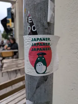 Street sticker The sticker features a round, white background with a prominent red circle in the center, reminiscent of the Japanese flag. Below the red circle, the character Totoro from Studio Ghibli is depicted in green and white. Totoro is holding a black umbrella over its head. Above Totoro and the red circle, the text "JAPANER, JAPANER, JAPANER!" is printed in a dark green, bold font, arranged in three lines.