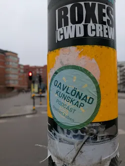Street sticker A round sticker featuring the text "Oavlönad Kunskap PODCAST @galleri.kuben" is affixed to a pole. The sticker has a light blue and green background with yellow accents.