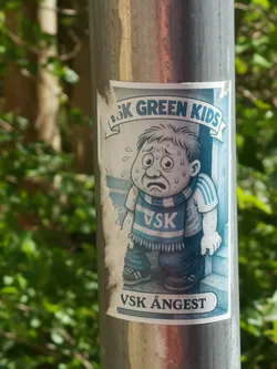 Street sticker A rectangular sticker affixed to a metallic pole, featuring a cartoon illustration of a young boy. The boy appears highly distressed, with wide, worried eyes, a downturned mouth, and visible sweat droplets on his face and arms. He has short, dark hair and is wearing a light-colored t-shirt with blue stripes on the sleeves, and a blue and white striped scarf around his neck that reads 'VSK'. His pants are dark blue, and he wears dark shoes. The background is a light blue, with a faint outline suggesting a doorway or corner. Above the boy, a banner reads 'SK GREEN KIDS' (the 'V' is partially obscured). Below the boy, another banner clearly states 'VSK ÅNGEST'. The overall color palette is dominated by cool tones of blue, white, and grey. The sticker shows signs of wear, particularly along its left edge, where it is slightly torn and faded.