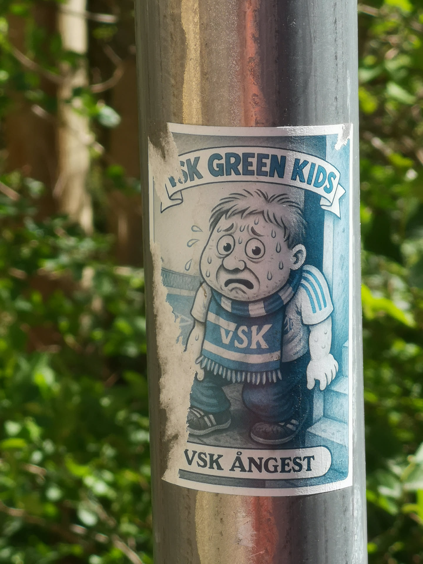 A rectangular sticker affixed to a metallic pole, featuring a cartoon illustration of a young boy. The boy appears highly distressed, with wide, worried eyes, a downturned mouth, and visible sweat droplets on his face and arms. He has short, dark hair and is wearing a light-colored t-shirt with blue stripes on the sleeves, and a blue and white striped scarf around his neck that reads 'VSK'. His pants are dark blue, and he wears dark shoes. The background is a light blue, with a faint outline suggesting a doorway or corner. Above the boy, a banner reads 'SK GREEN KIDS' (the 'V' is partially obscured). Below the boy, another banner clearly states 'VSK ÅNGEST'. The overall color palette is dominated by cool tones of blue, white, and grey. The sticker shows signs of wear, particularly along its left edge, where it is slightly torn and faded.