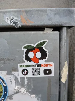 Street sticker A vibrant, die-cut sticker featuring a stylized, anthropomorphic mango character. The mango itself is a bright orange, forming the face, adorned with large, round white glasses with thin orange frames. A mass of black, curly hair frames the face, topped with two dark green leaves. Below the character, a white rectangular banner displays the text 'MANGOINTHENORTH', with 'MANGOIN' in green and 'THENORTH' in orange. To the left of the text, there's a black TikTok logo, and to the right, a black YouTube play button logo. In the center, below the text, a clear black and white QR code is present.