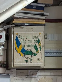 Street sticker A large, rectangular sticker with a cream-colored background, showing signs of wear and peeling at the edges. It features a cartoon illustration of a supporter from behind, wearing a green shirt and white shorts, holding two crossed flags: the Swedish flag (blue and yellow) on the left, and a green and white striped flag with a yellow 'H' (Hammarby) on the right. Above the illustration, the text 'Jag vill leva Jag vill dö på söder.' is written in a green gothic-style font.