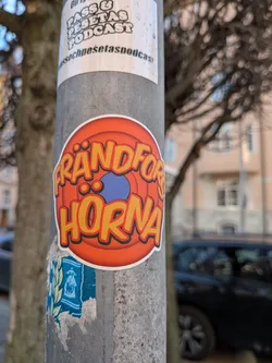 Street sticker A circular sticker featuring bold, stylized text that reads 'FRÄNDFÖR HORNA'. The letters are primarily yellow with an orange outline and a subtle black drop shadow, giving them a three-dimensional, playful appearance. The background of the sticker consists of concentric circles in shades of orange, fading from a brighter orange at the center to a darker, more reddish-orange towards the edge, reminiscent of classic animation studio logos. A small, solid blue oval shape is visible in the center, partially obscured by the text. The entire design is bordered by a clean white outline, making it pop against the textured grey pole.