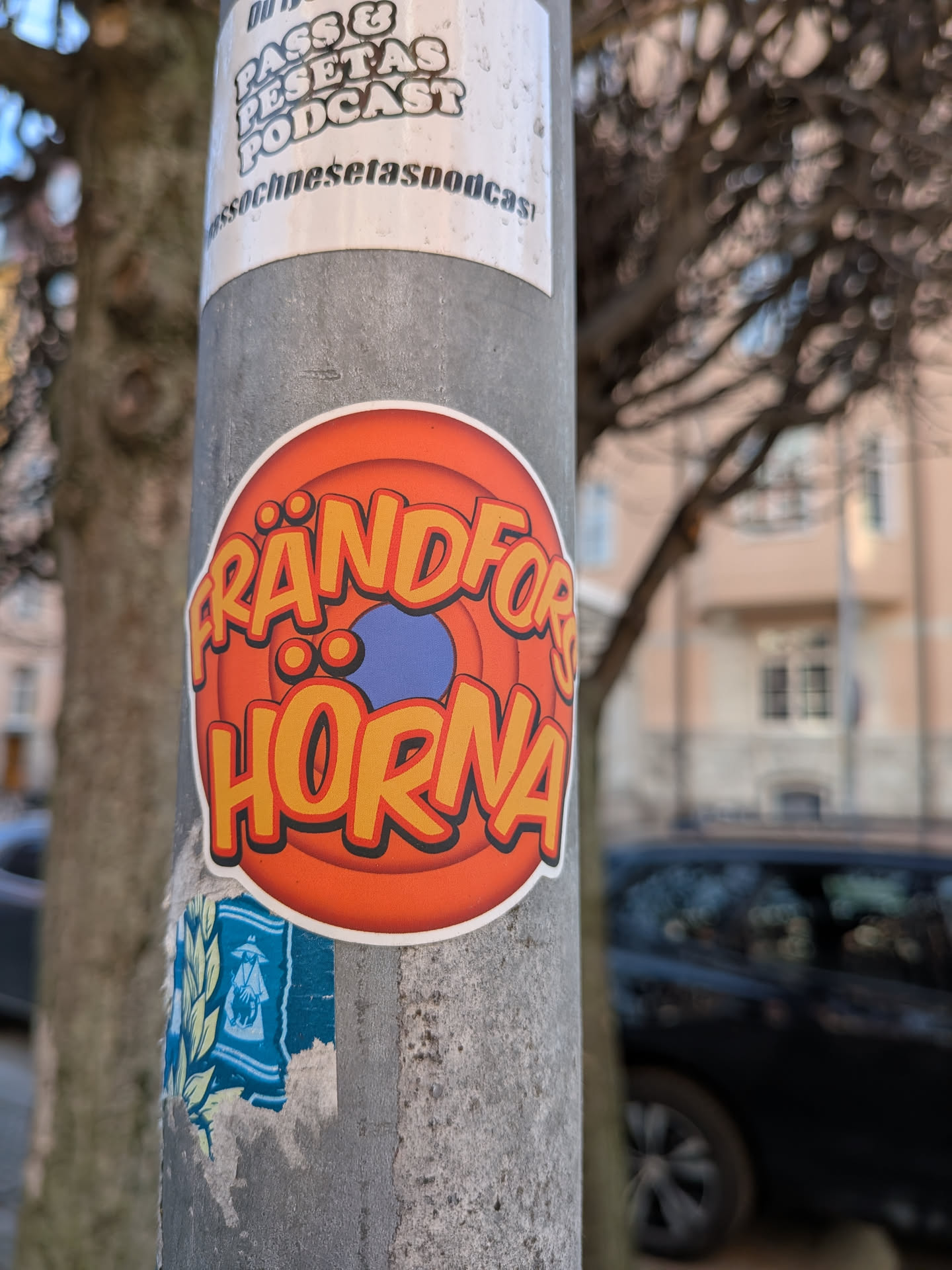 A circular sticker featuring bold, stylized text that reads 'FRÄNDFÖR HORNA'. The letters are primarily yellow with an orange outline and a subtle black drop shadow, giving them a three-dimensional, playful appearance. The background of the sticker consists of concentric circles in shades of orange, fading from a brighter orange at the center to a darker, more reddish-orange towards the edge, reminiscent of classic animation studio logos. A small, solid blue oval shape is visible in the center, partially obscured by the text. The entire design is bordered by a clean white outline, making it pop against the textured grey pole.