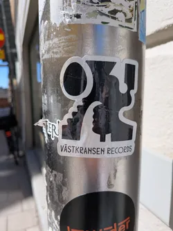 Street sticker A black and white sticker featuring a bold, abstract graphic that appears to be a stylized letter combination or two opposing profiles, enclosed by a thick white outline. The main graphic is black with a white border. Below this main graphic, a white rectangular banner contains the text 'VÄSTKRANSEN RECORDS' in black, sans-serif capital letters. The sticker shows signs of wear, including wrinkles and slight peeling, indicating it's a physical sticker on an outdoor pole.