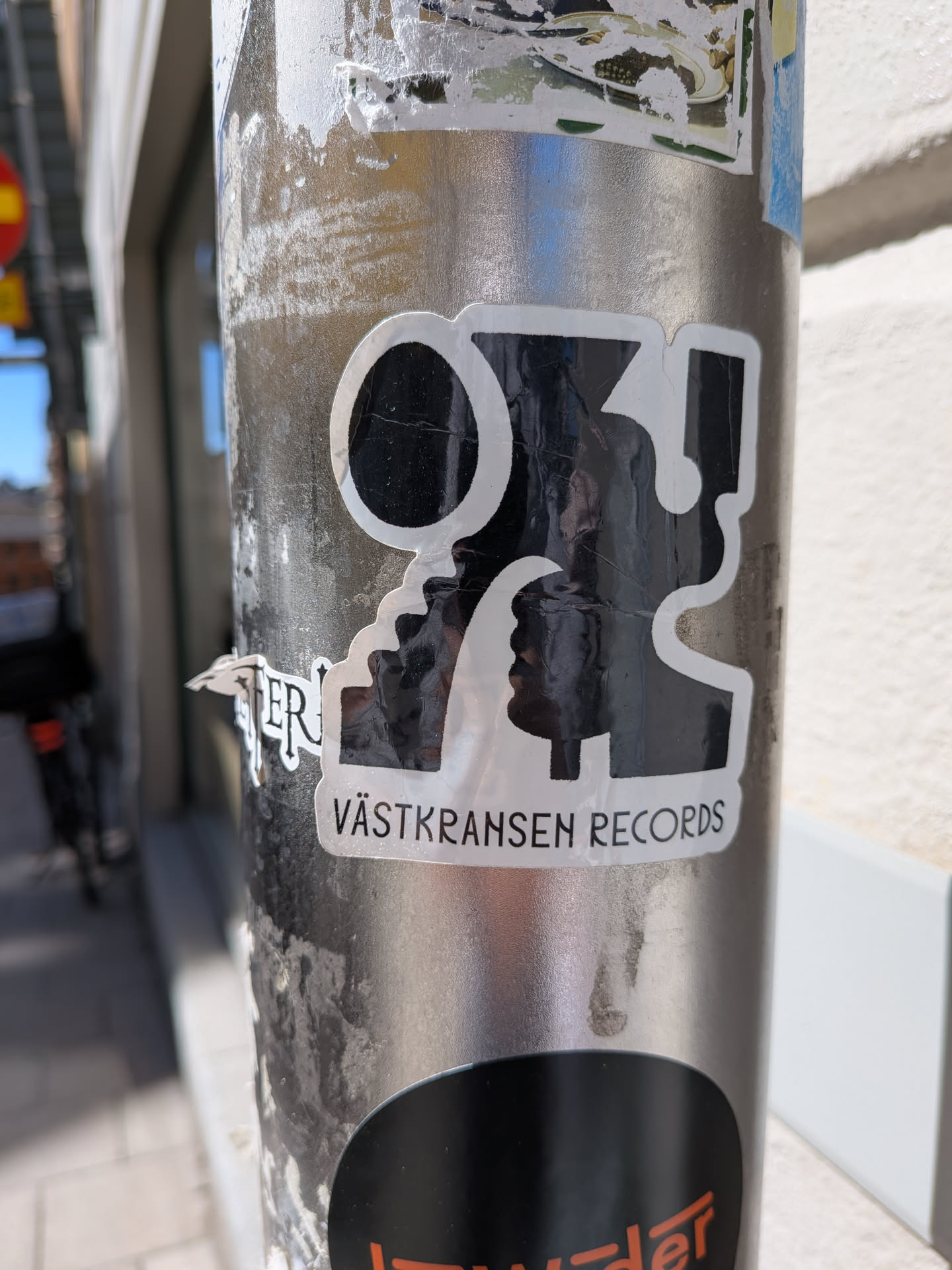 A black and white sticker featuring a bold, abstract graphic that appears to be a stylized letter combination or two opposing profiles, enclosed by a thick white outline. The main graphic is black with a white border. Below this main graphic, a white rectangular banner contains the text 'VÄSTKRANSEN RECORDS' in black, sans-serif capital letters. The sticker shows signs of wear, including wrinkles and slight peeling, indicating it's a physical sticker on an outdoor pole.